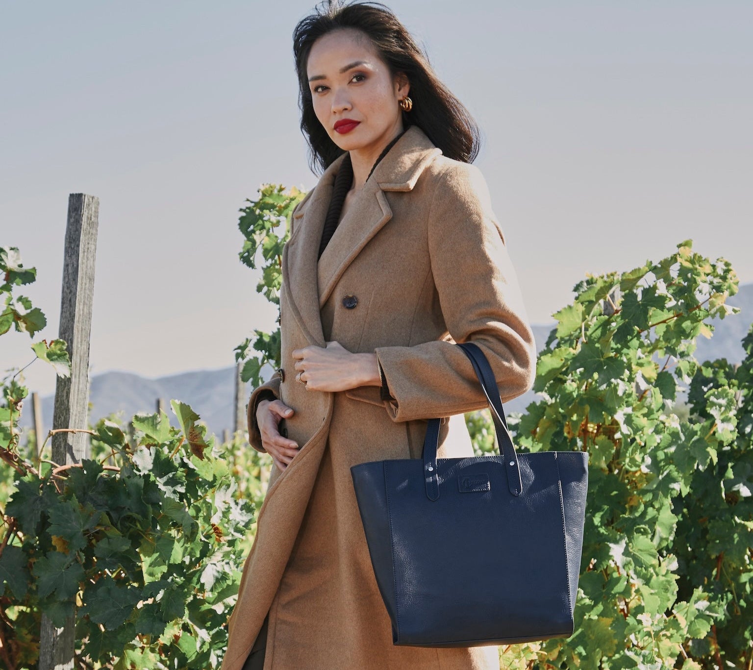 Woman in a beige coat holding a navy blue handbag in a vineyard setting